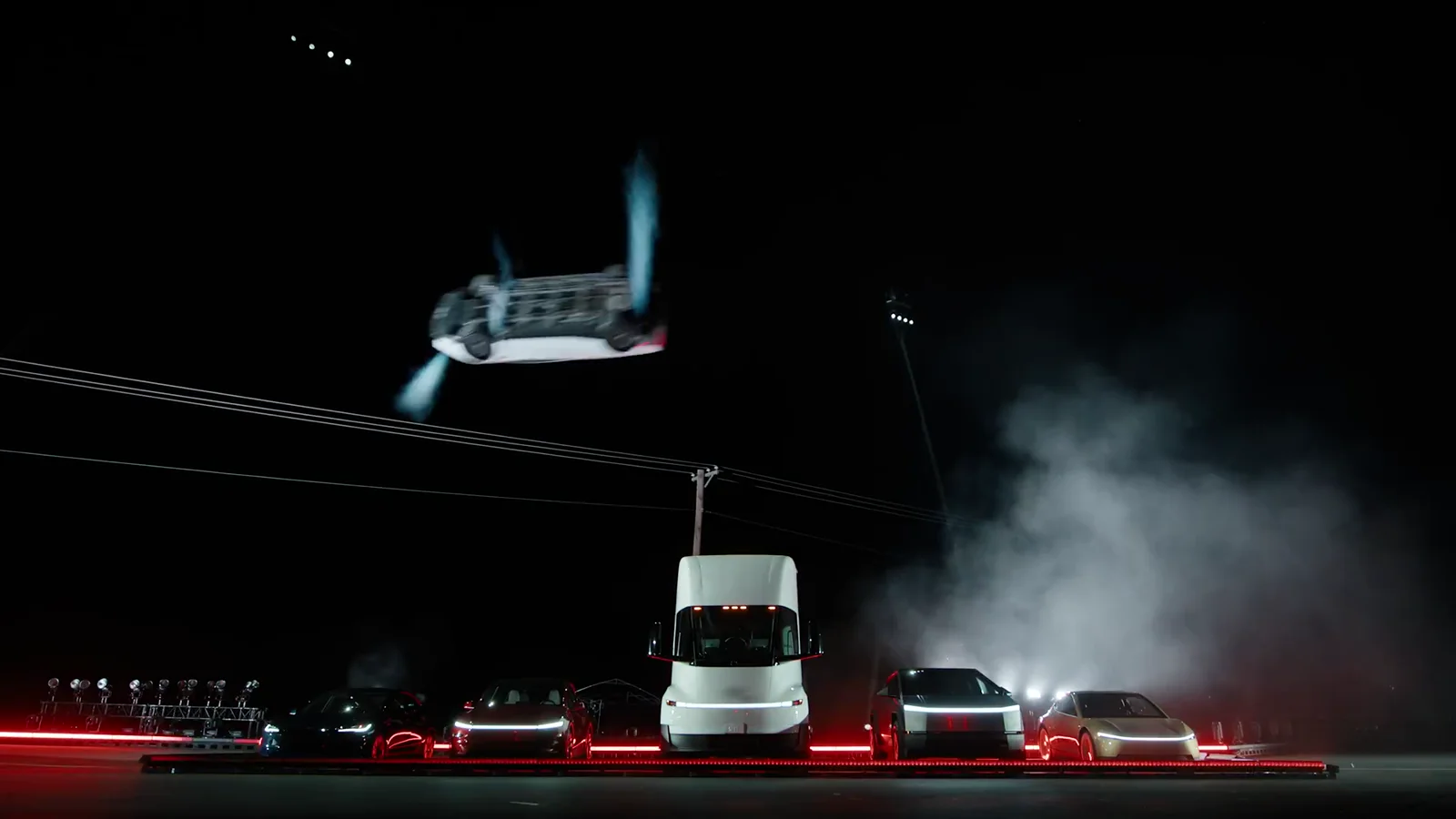 Tesla's Roadster upside down in mid-air above a line of parked Teslas, thrusters firing.
